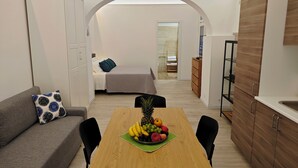 Ein kompaktes Hotelzimmer mit Bett, einer kleinen Küchenzeile, einem Essbereich mit Obstschale und einem grauen Sofa.