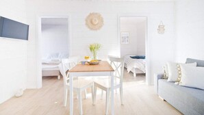 A bright, white interior with a wooden dining table, white chairs, a grey sofa, and a bed with white bedding.