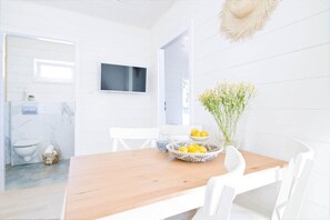 A bright, white interior with a wooden dining table, white chairs, a TV mounted on the wall, and a vase with yellow flowers.