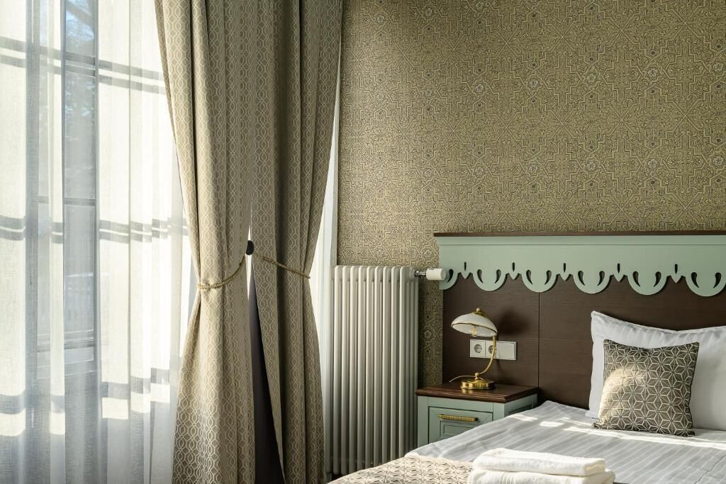 Una camera d'albergo con un letto, un comodino, una lampada, un termosifone e una finestra con tende.