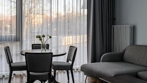 A modern living room with a grey sofa, a dining table with four chairs, and a large window with sheer curtains.