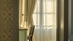 A room with a patterned wallpaper, a wooden floor, a chair, and a desk with a chair.