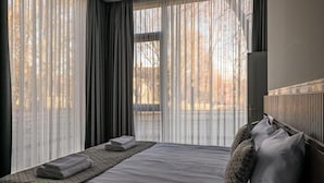 A hotel room with a bed, a bedside table with towels, and a view of trees through sheer curtains.