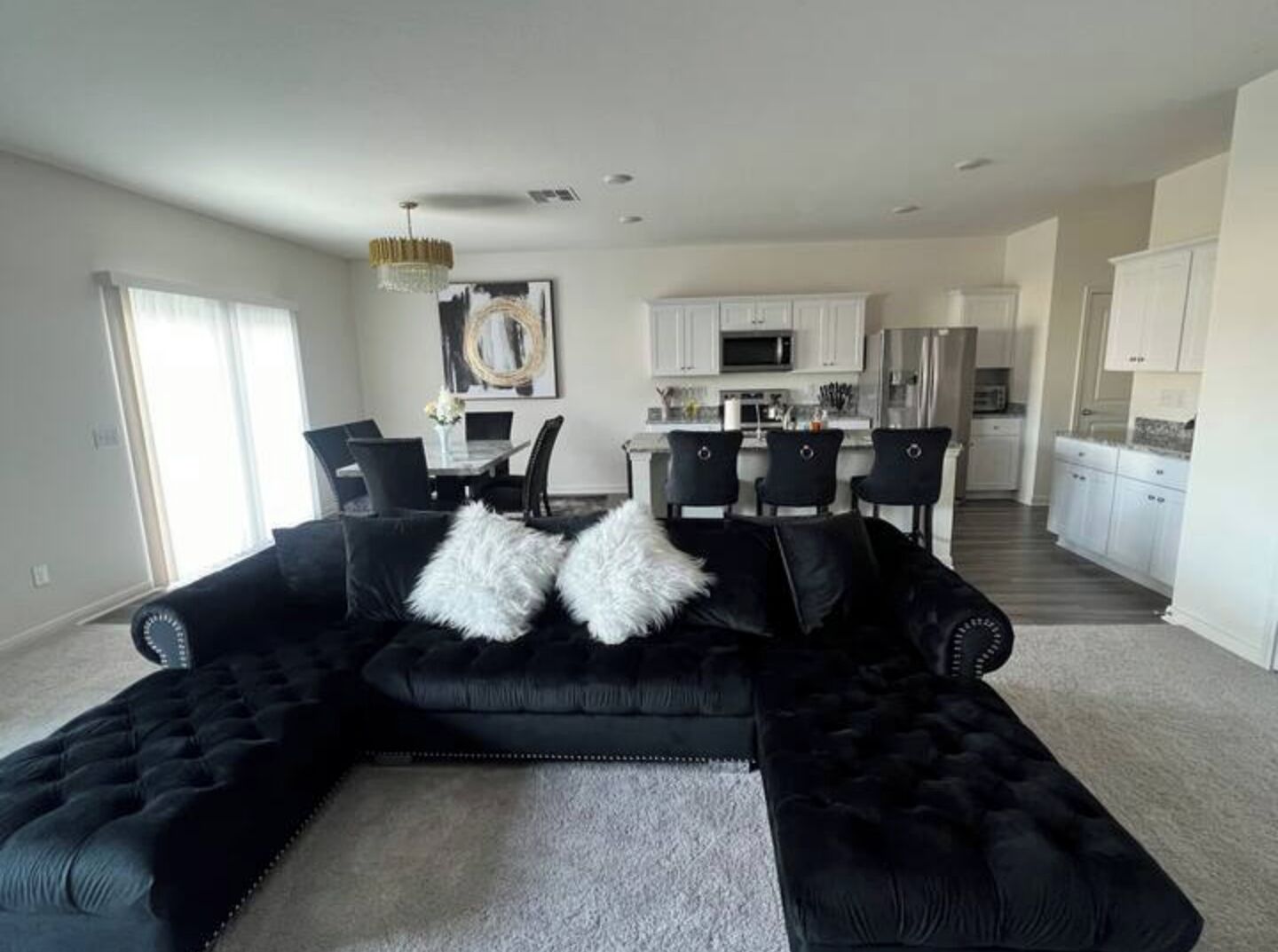 A modern living room with a black sectional sofa, a dining area with a chandelier, and a kitchen in the background.