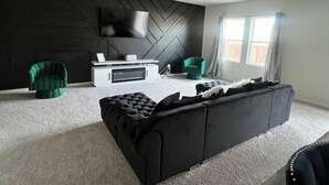 Een moderne woonkamer met een zwart modulaire bank, twee groene fauteuils, een witte tv-meubel en een groot raam met gordijnen.