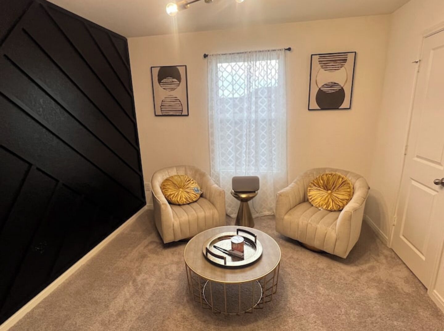 Een kamer met twee beige fauteuils, een ronde salontafel en een bijzettafeltje. Twee ingelijste kunstwerken hangen aan de muur, een raam met gordijnen met een patroon, en een deur.