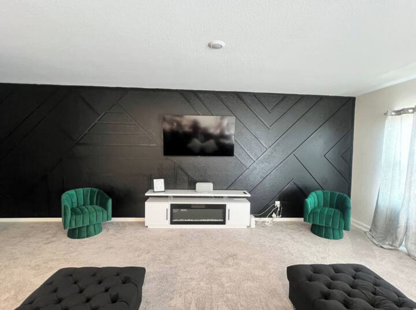 A modern living room with a black herringbone-patterned wall, a flat-screen TV mounted above a white entertainment unit, and two green armchairs.
