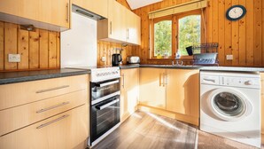 A kitchen with wooden cabinets, a stove, an oven, a washing machine, and a window with a view of trees.