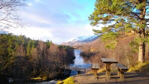 A scenic riverside view with a wooden bridge, picnic tables, and a mountainous backdrop.