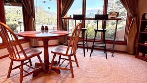 Un coin repas avec une table ronde et quatre chaises en bois, de grandes fenêtres avec des rideaux et une vue sur un paysage montagneux.