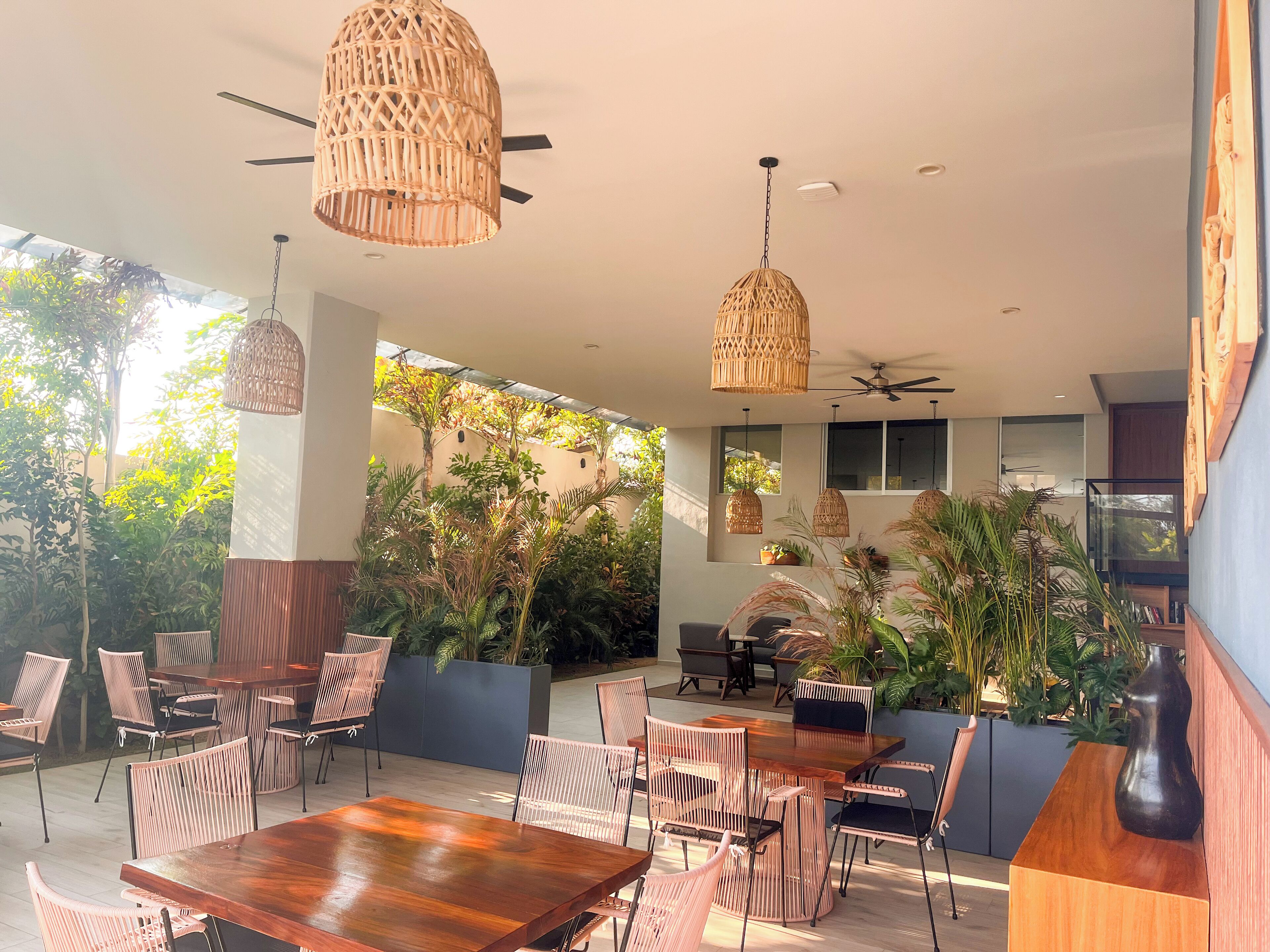 An outdoor dining area with wooden tables and wicker chairs, surrounded by lush greenery and potted plants.