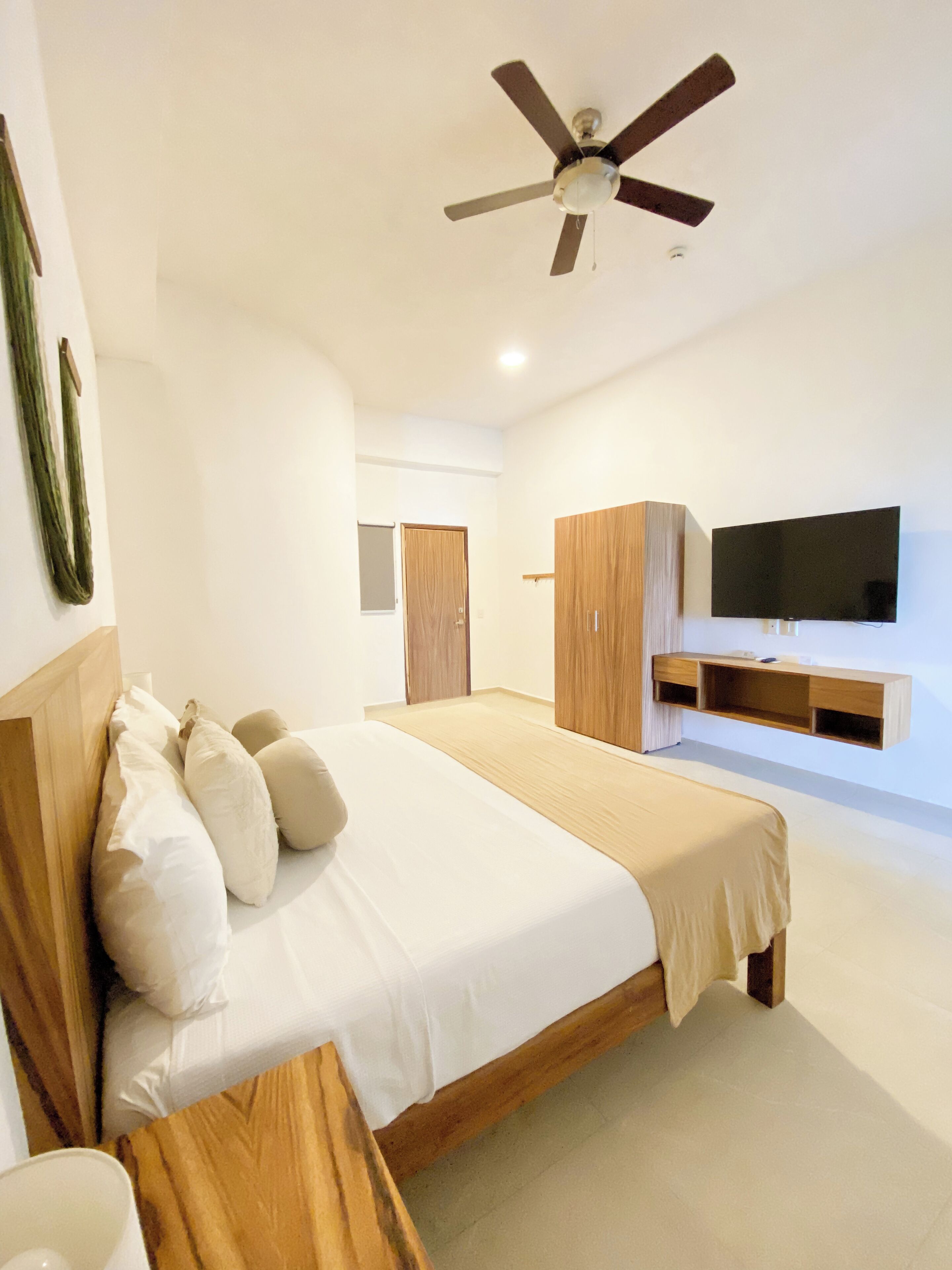 A modern bedroom with a large bed, wooden headboard, a ceiling fan, a TV mounted on the wall, and a wooden wardrobe.