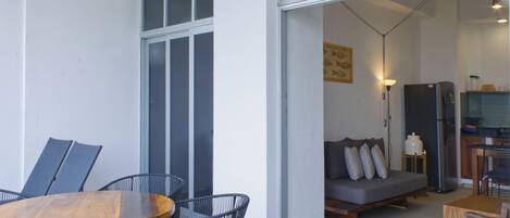 An open-plan living and dining area with a wooden dining table, black metal chairs, a grey sofa, and a kitchen visible through an open doorway.