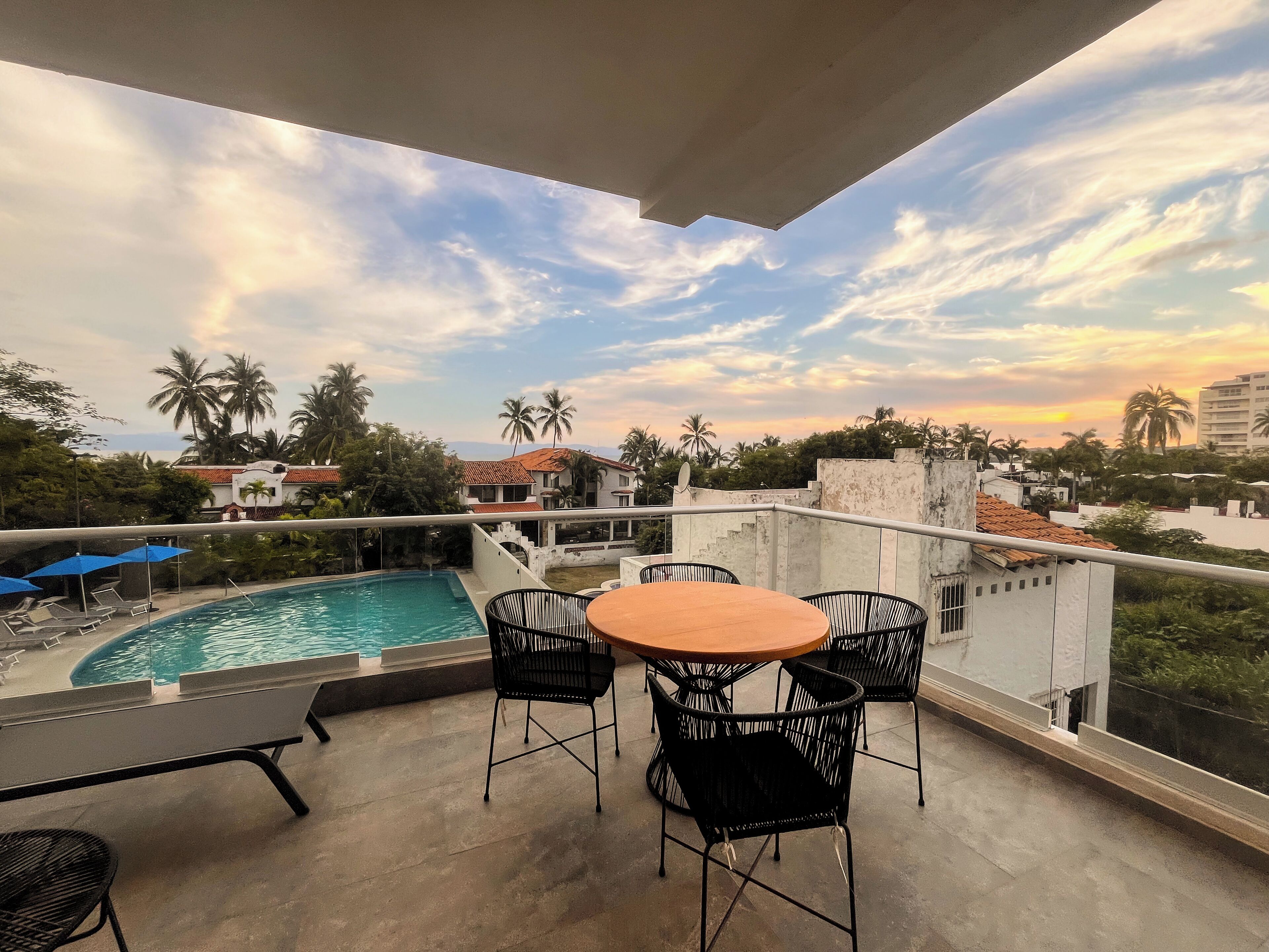 Un balcón con piscina, sillas y una mesa, con vistas a una zona residencial con palmeras y casas.