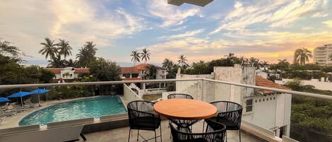 Un balcón con piscina, sillas y una mesa, con vistas a una zona residencial con palmeras y casas.