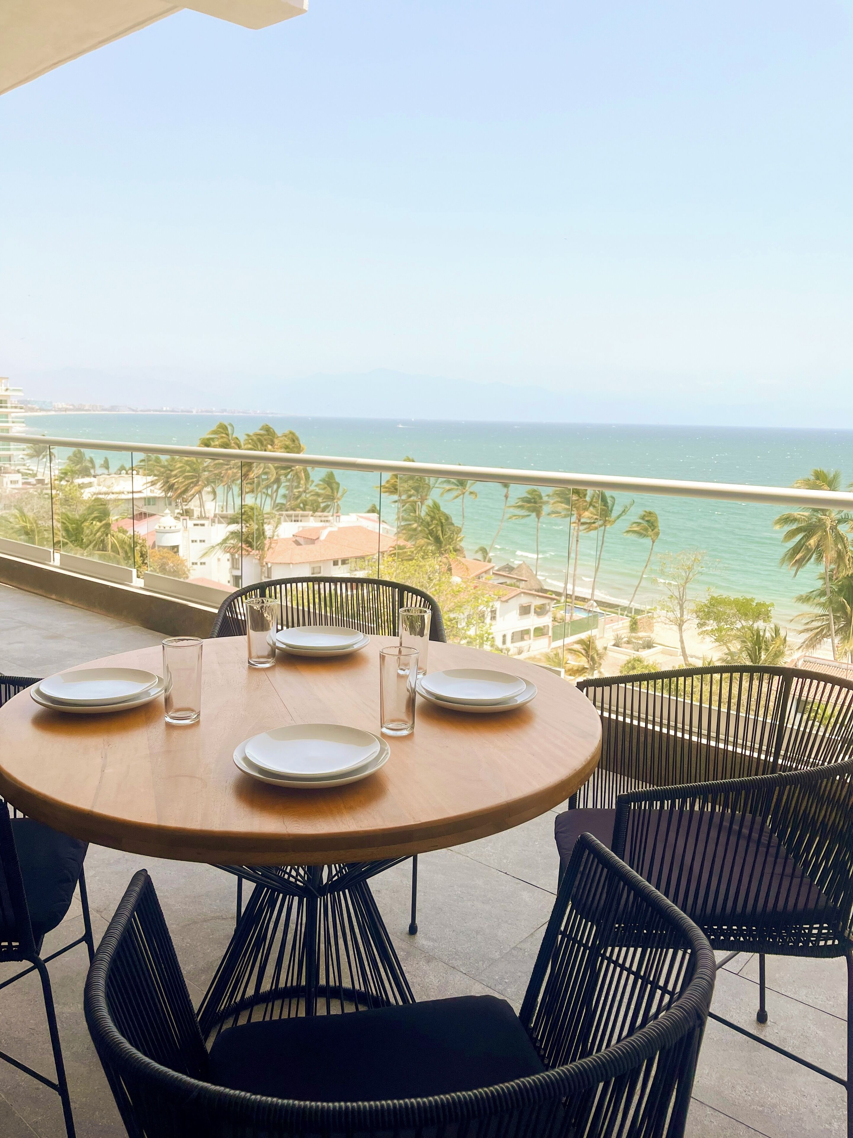 A round table with four plates and glasses on a balcony overlooking a beach with palm trees and a clear sky.