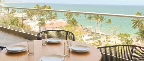 A round table with four plates and glasses on a balcony overlooking a beach with palm trees and a clear sky.