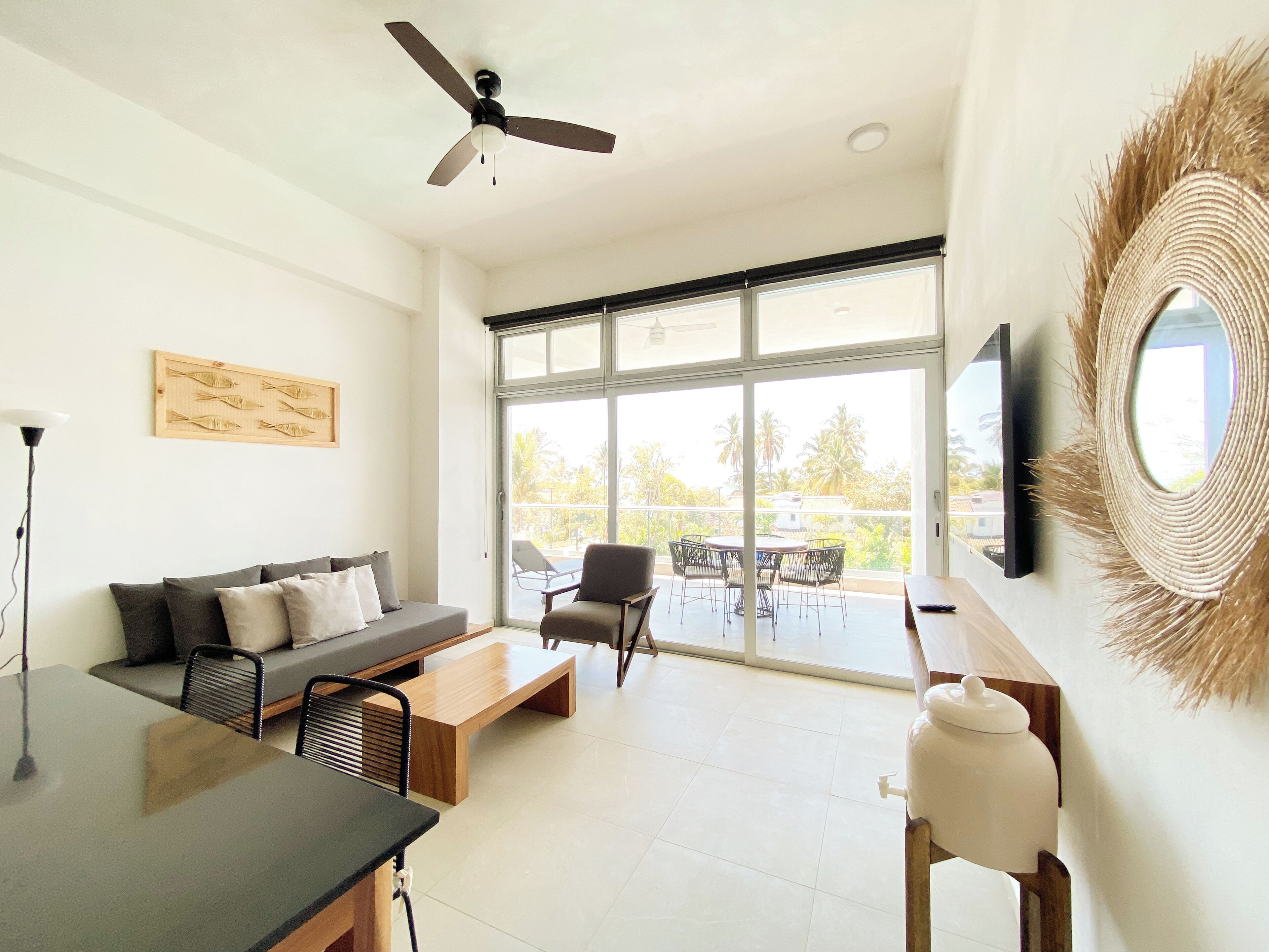 Uma sala de estar moderna com sofá, mesa de centro e cadeiras, ventilador de teto, TV fixada na parede e grandes janelas com vista para o exterior.