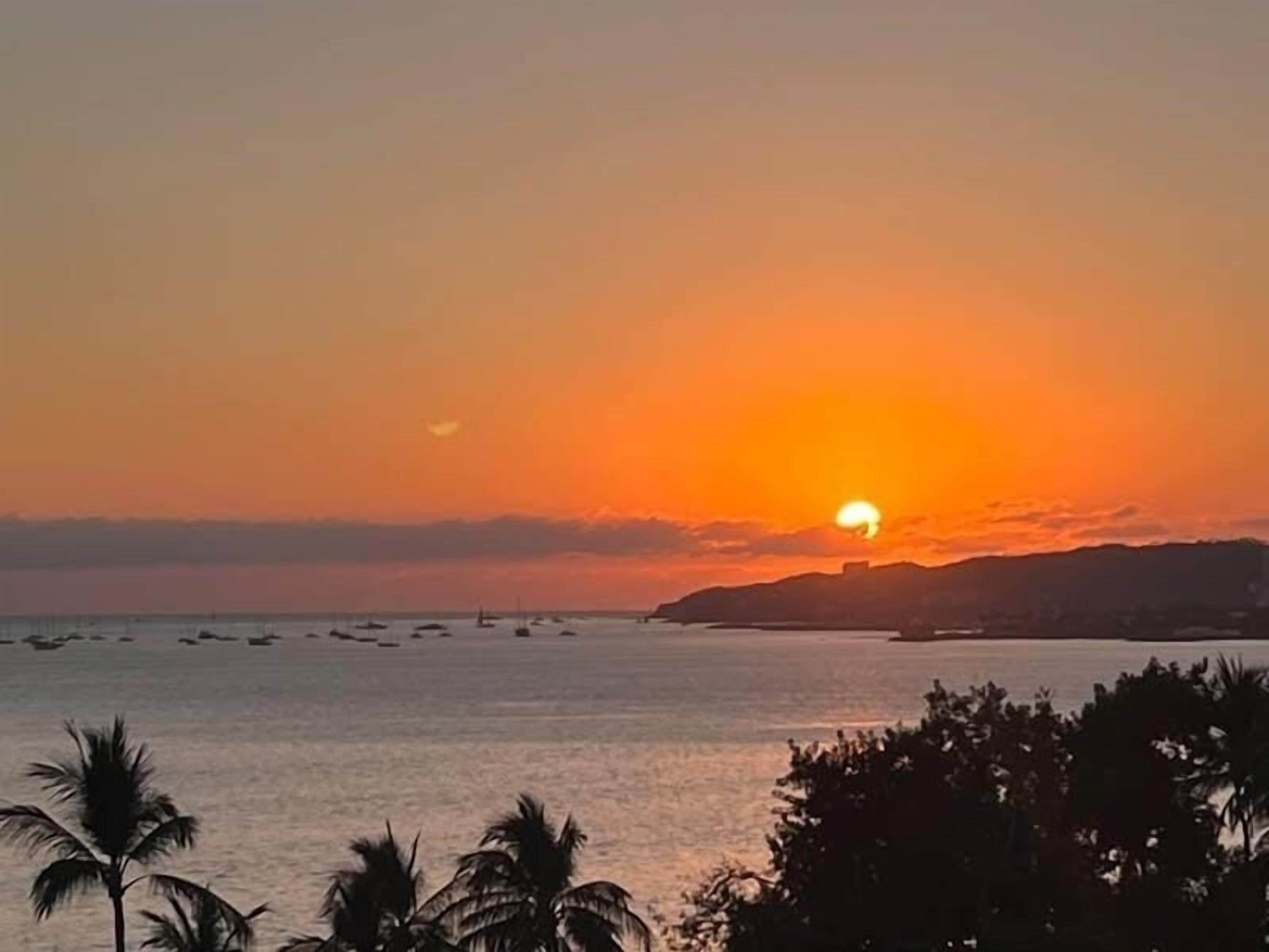 A sunset over a calm sea with boats, palm trees, and a distant shoreline.