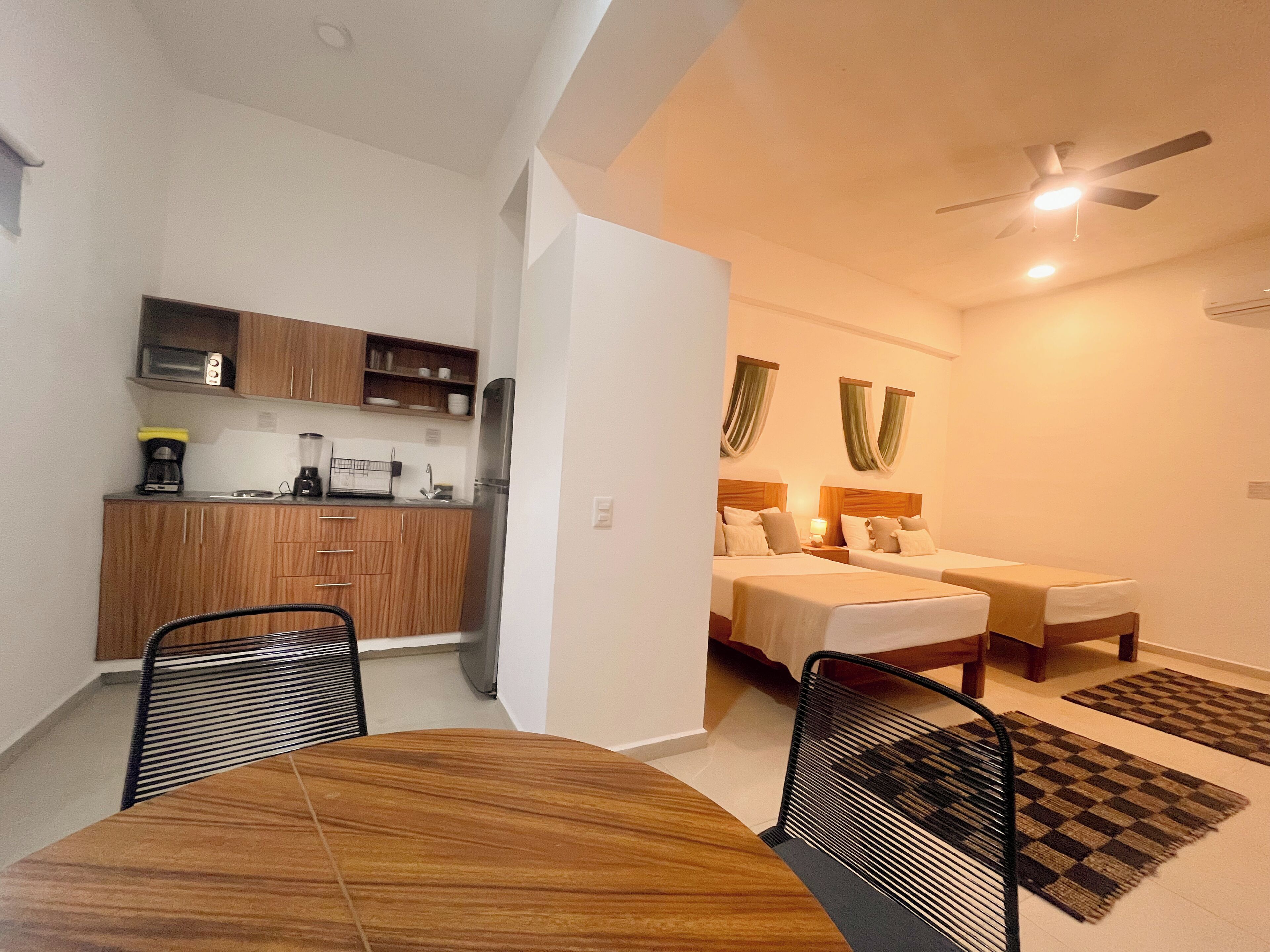 Une chambre d’hôtel compacte comprenant un coin cuisine, deux lits, une table à manger et un ventilateur de plafond.