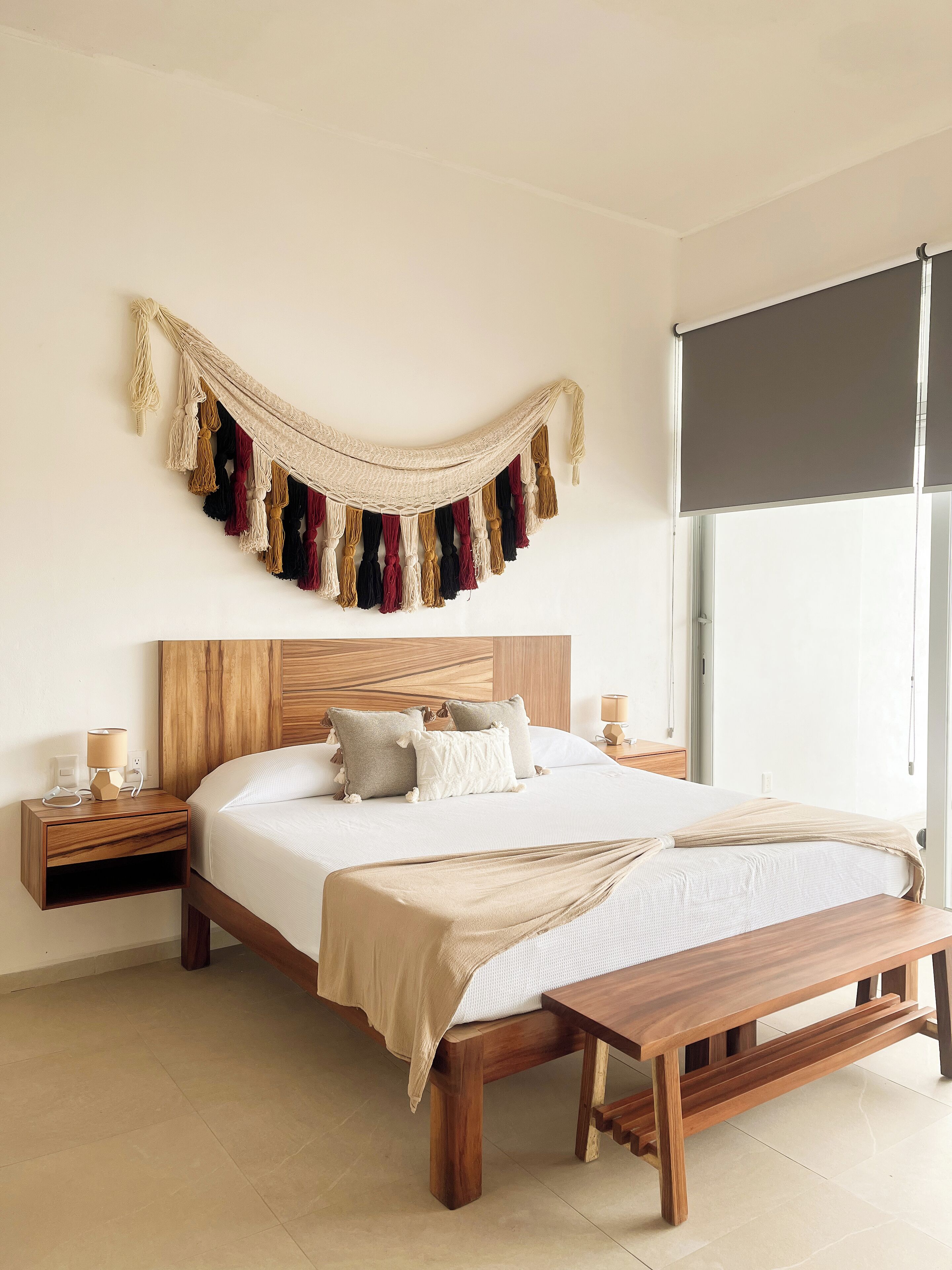 A bedroom with a wooden bed, a wooden nightstand, a bedside table, a bench, a wall-mounted macrame decoration, and a window with a blind.