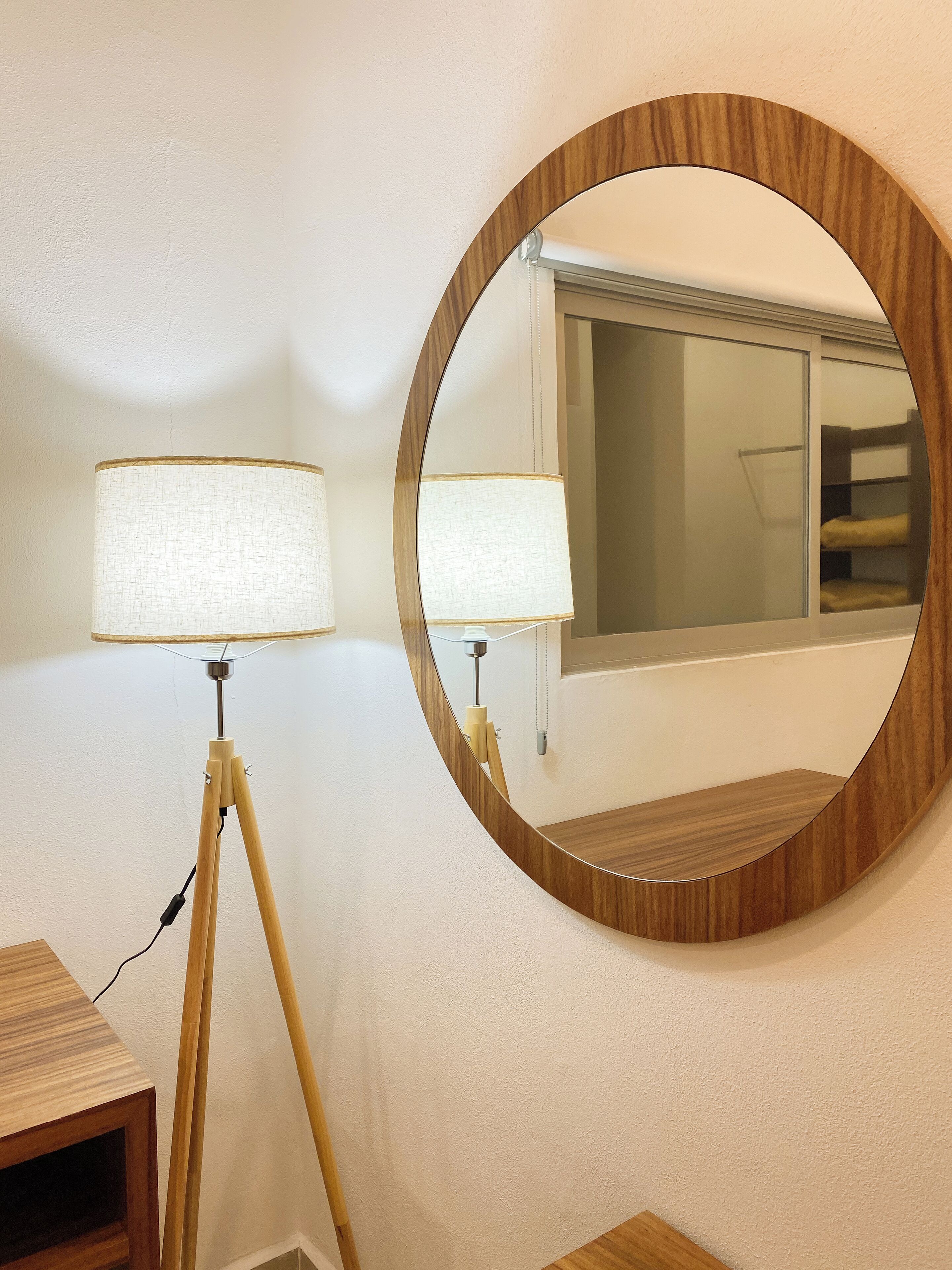 A round mirror with a wooden frame on a wall, reflecting a window and a bed with a white headboard.