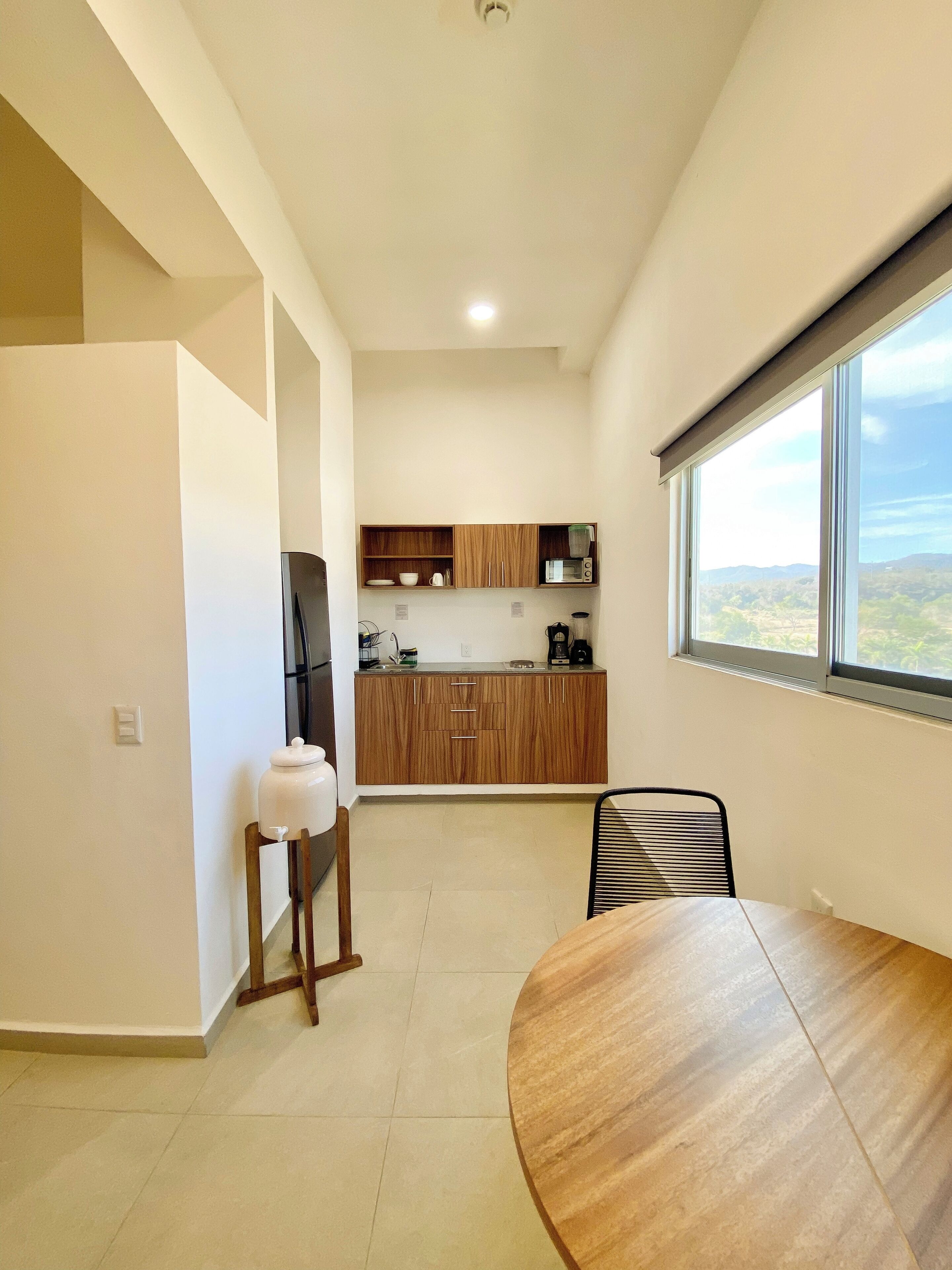A modern kitchen with wooden cabinets, a microwave, and a coffee maker. There is a dining table with chairs and a view of a mountainous landscape through a large window.
