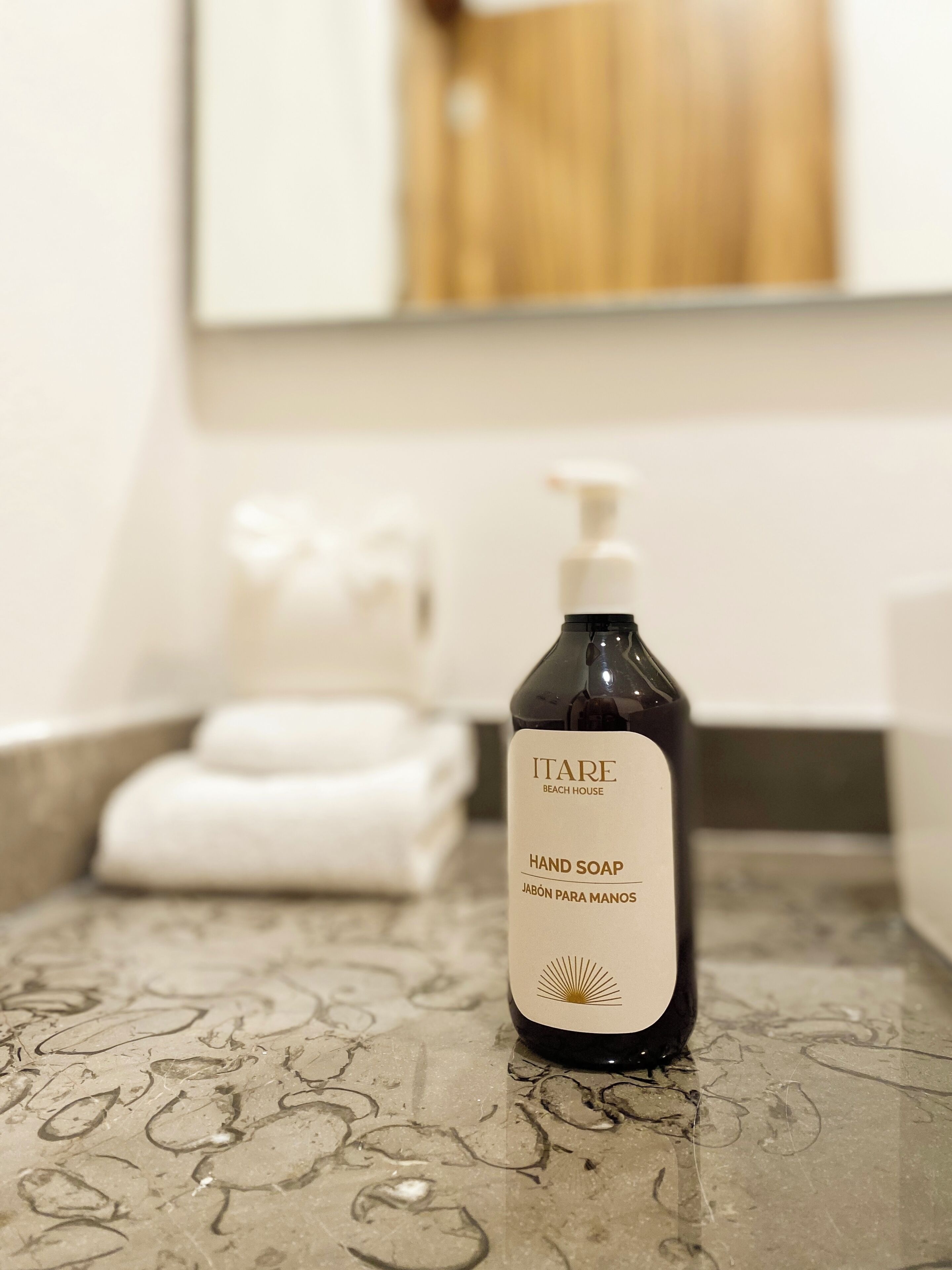 A bottle of Itare Beach House hand soap on a bathroom counter with a towel and a mirror in the background.
