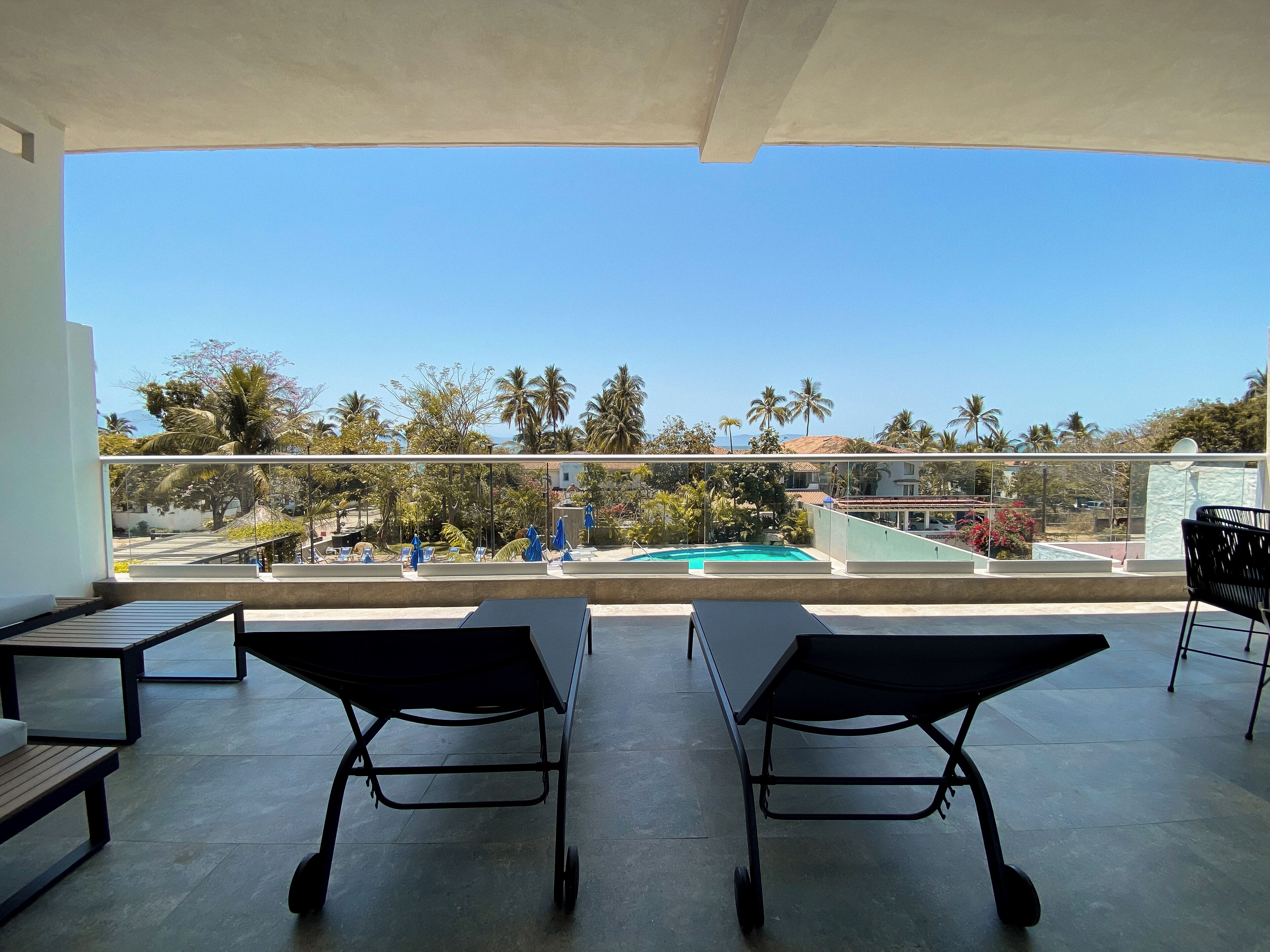 A balcony with two lounge chairs, a pool, and a view of palm trees and buildings.