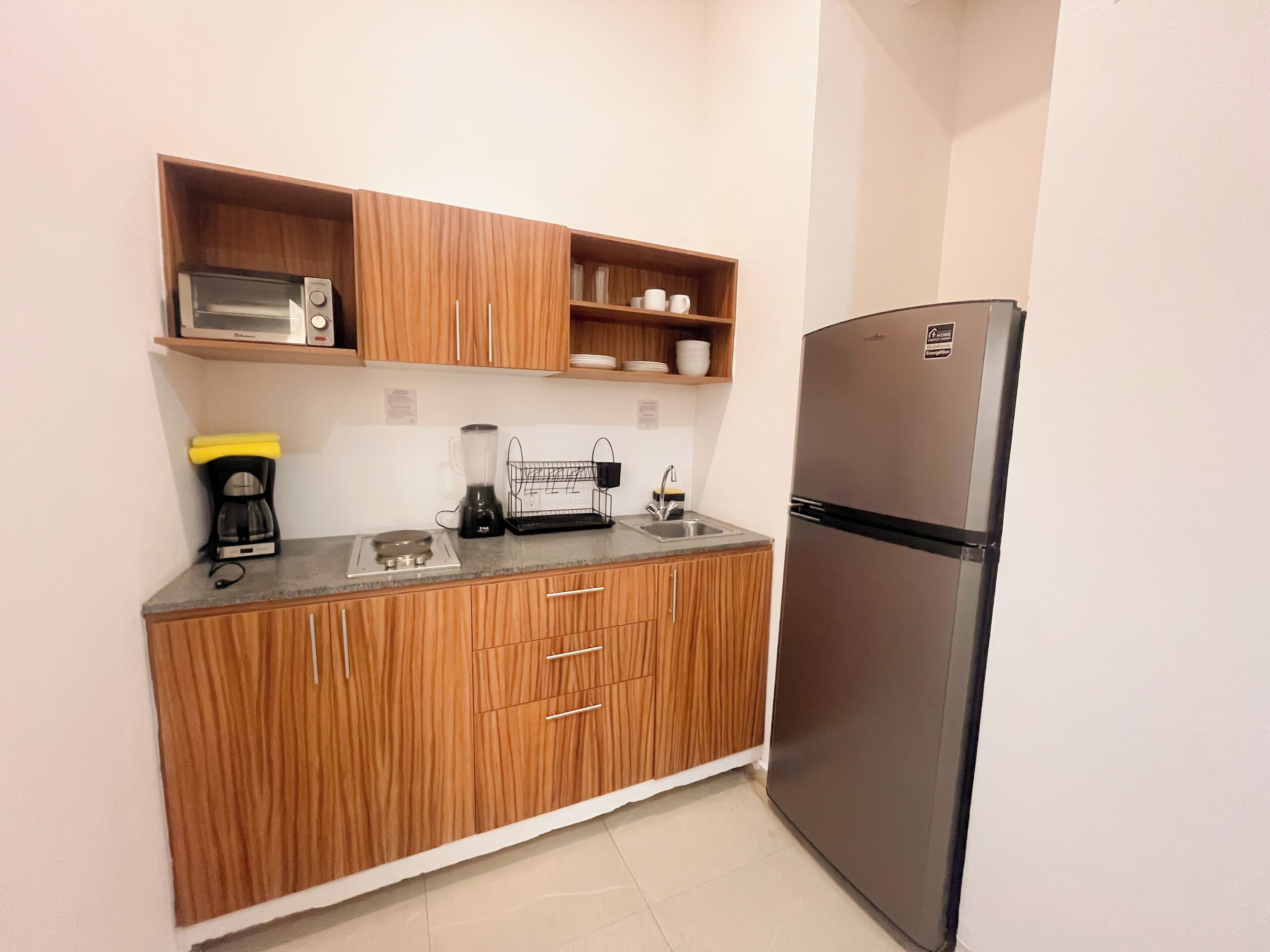 A compact kitchen with wooden cabinets, a stainless steel refrigerator, a coffee maker, and a blender on the countertop.