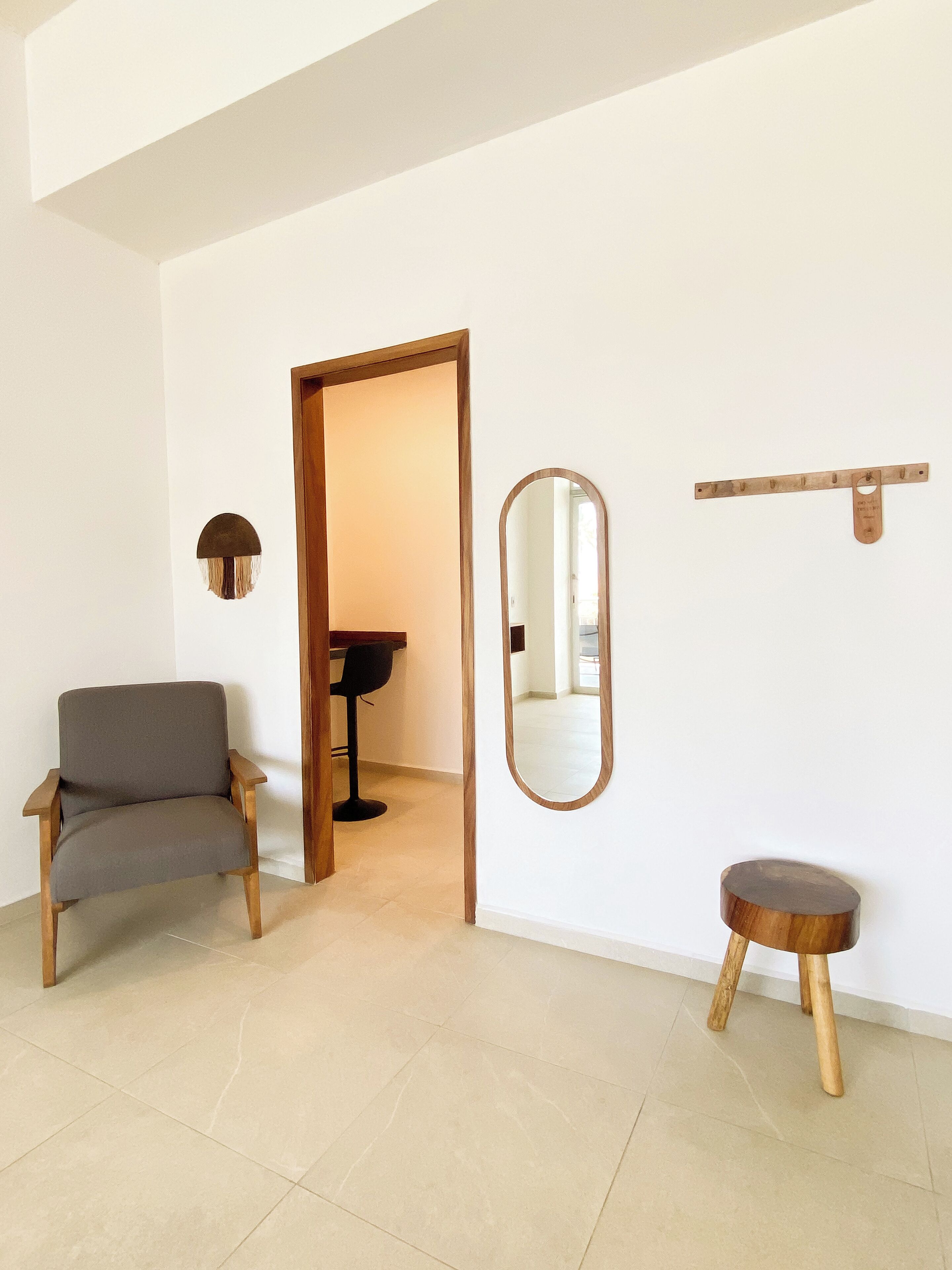 Un interior moderno con un sillón gris, un taburete de madera, un espejo y una puerta que da a otra habitación.