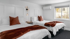 A hotel room with two beds, white bedding, brown throw blankets, a bedside table with a lamp, and an air conditioning unit. - Lincoln Boutique Motel- Lower Room #6 (Ellicottville)