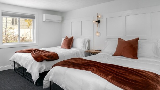 A hotel room with two beds, white bedding, brown throw blankets, a nightstand, and a wall-mounted light fixture.