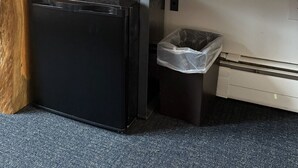 A compact hotel room with a wooden desk, a coffee maker, a trash bin, a black refrigerator, and a small trash bag.