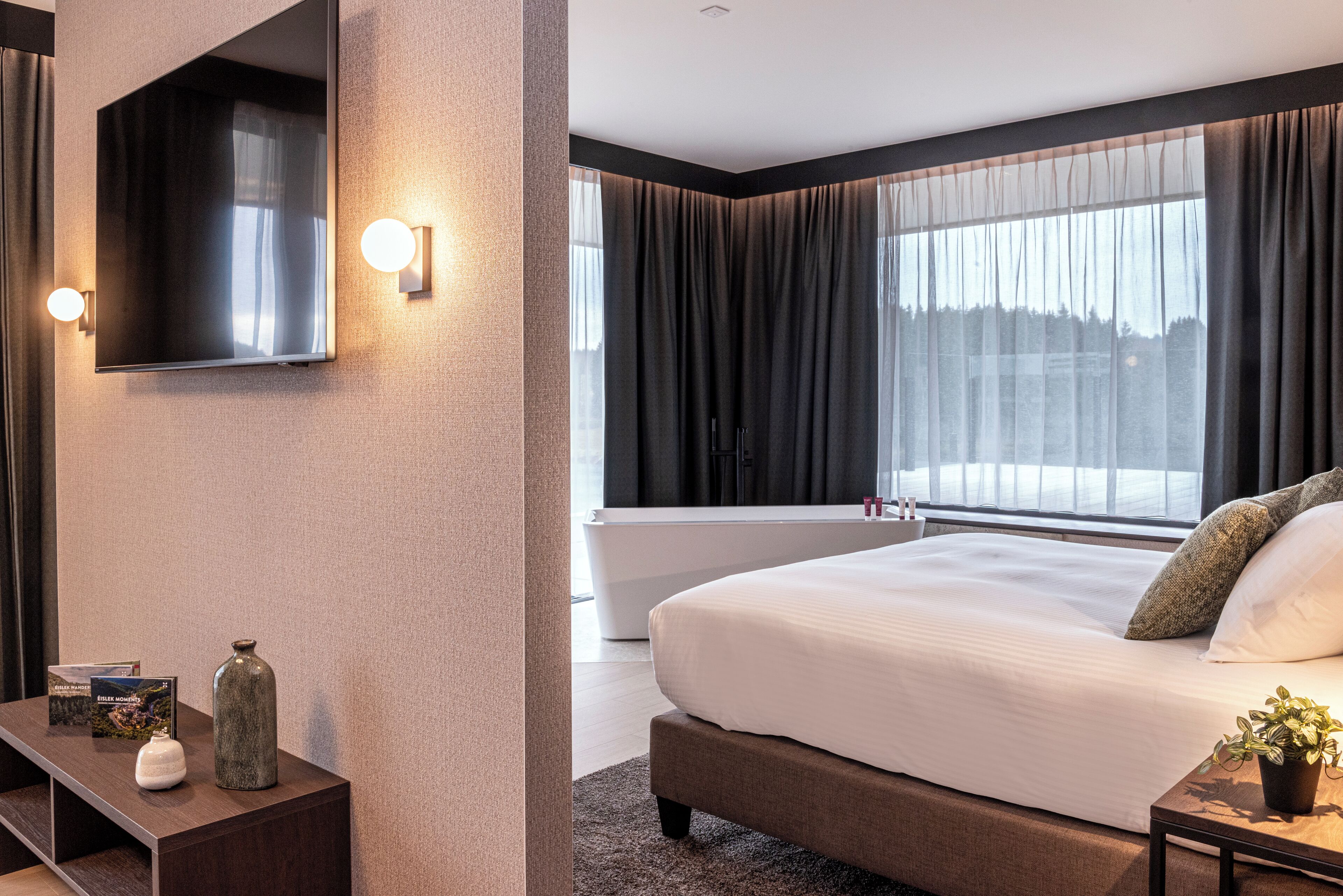 Ein modernes Hotelzimmer mit einem großen Bett, einem flachbildfernseher an der Wand, einer Badewanne und einem großen Fenster mit Vorhängen.