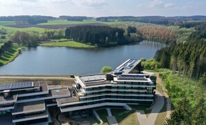 A modern building with a curved roof and multiple floors, surrounded by a natural landscape including a lake and forested hills.