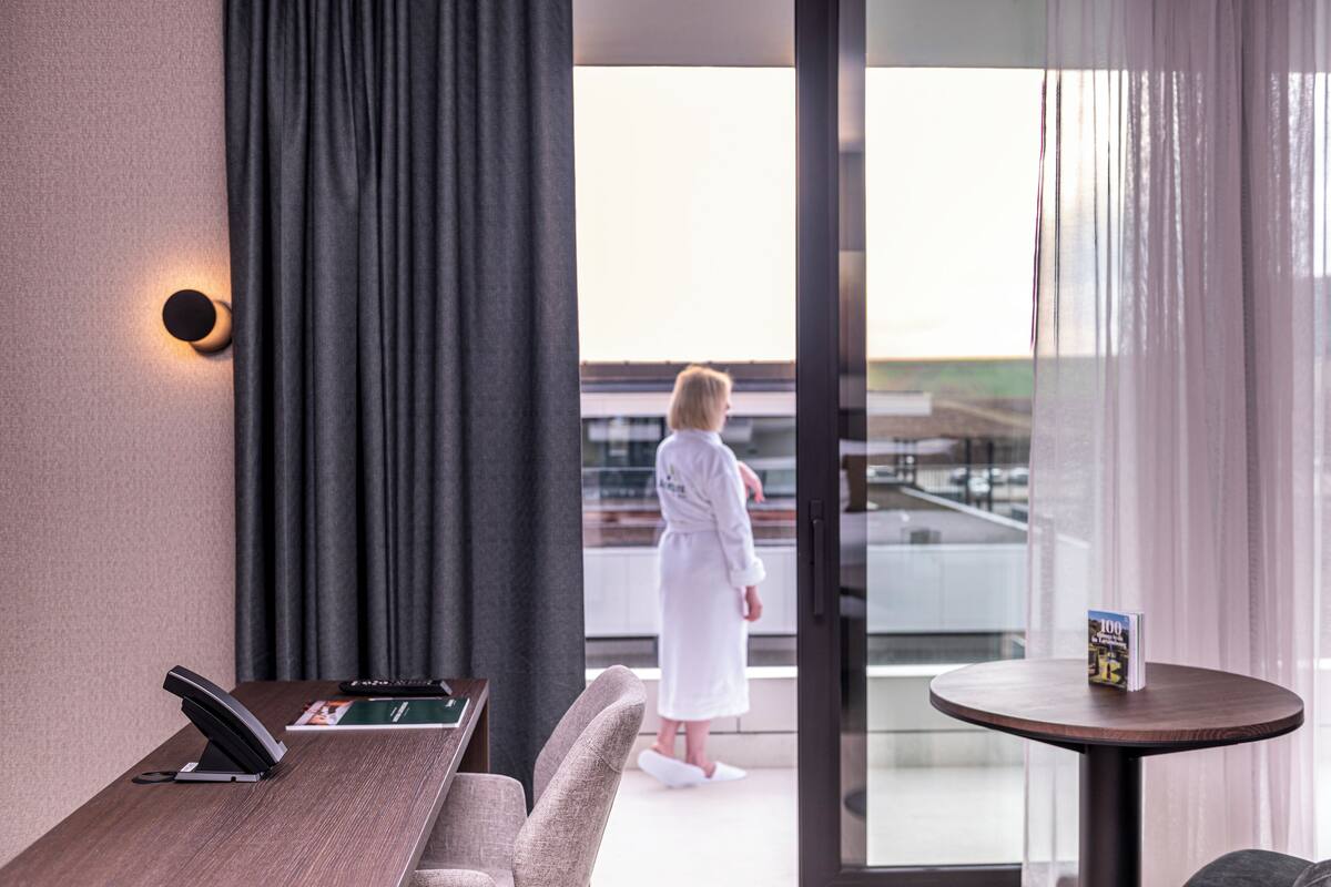Une chambre d’hôtel avec un bureau en bois, un téléphone, deux chaises, une petite table avec un livre, et une vue sur une personne en peignoir, visible à travers la fenêtre.