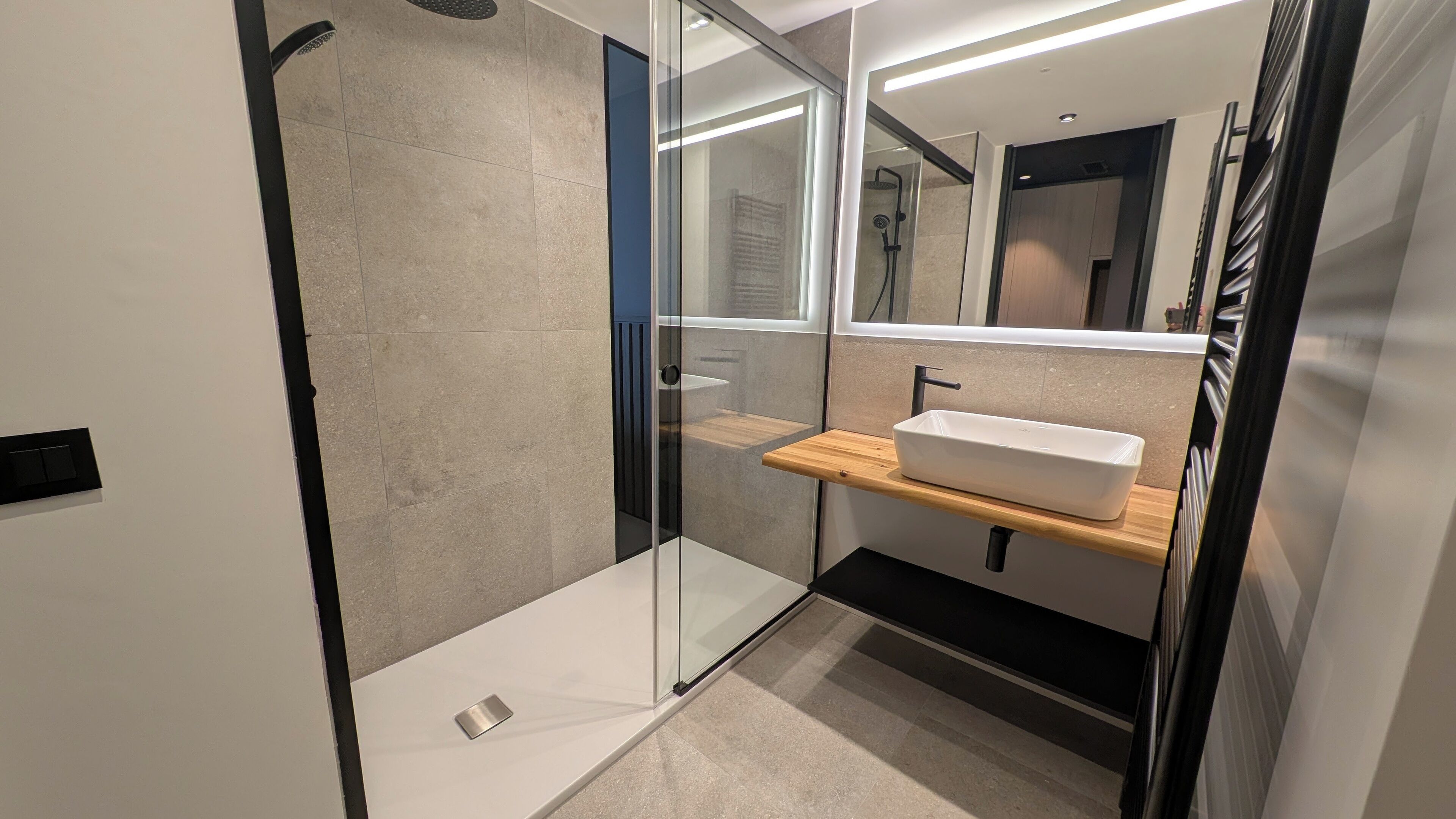 Een moderne badkamer met een douchecabine van glas, een witte wasbak op een houten plank en een spiegel erboven.