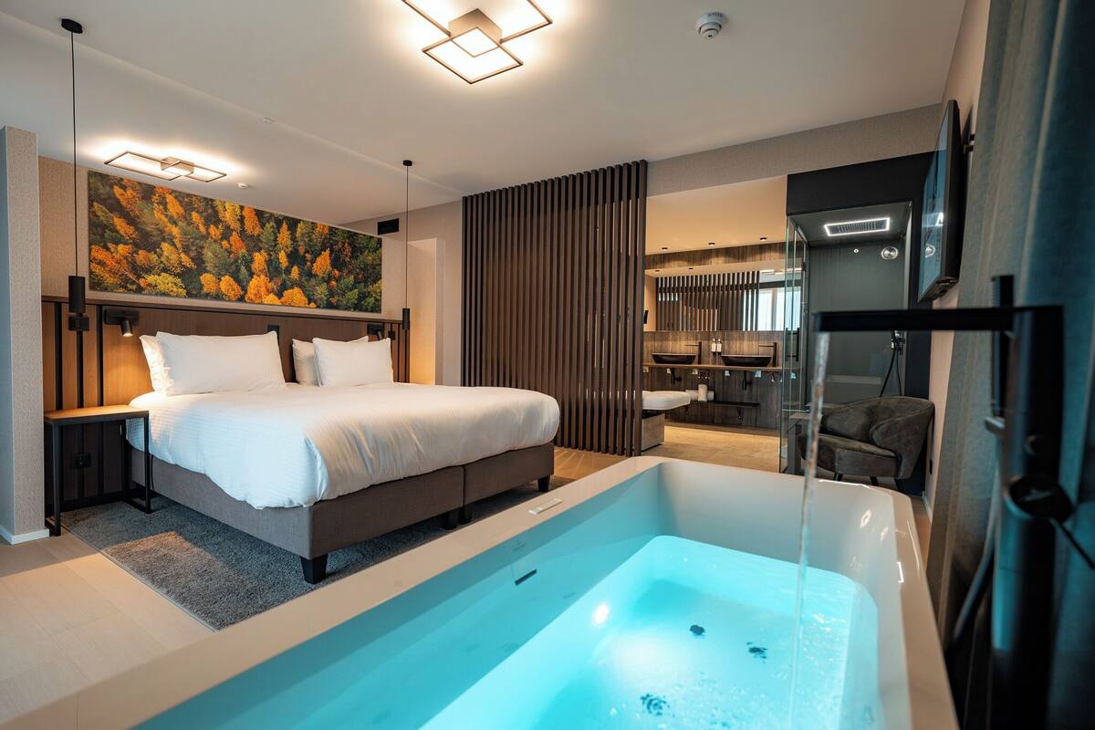 Une chambre d’hôtel moderne avec un grand lit, une baignoire, une salle de bain équipée d’un lavabo et d’un miroir, et un tableau accroché au mur.