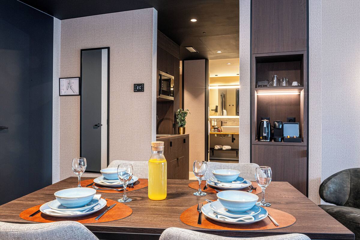 Un coin repas avec une table mise pour quatre personnes, comprenant des assiettes, des bols, des couverts et des verres. Une bouteille d’une boisson jaune est posée sur la table.