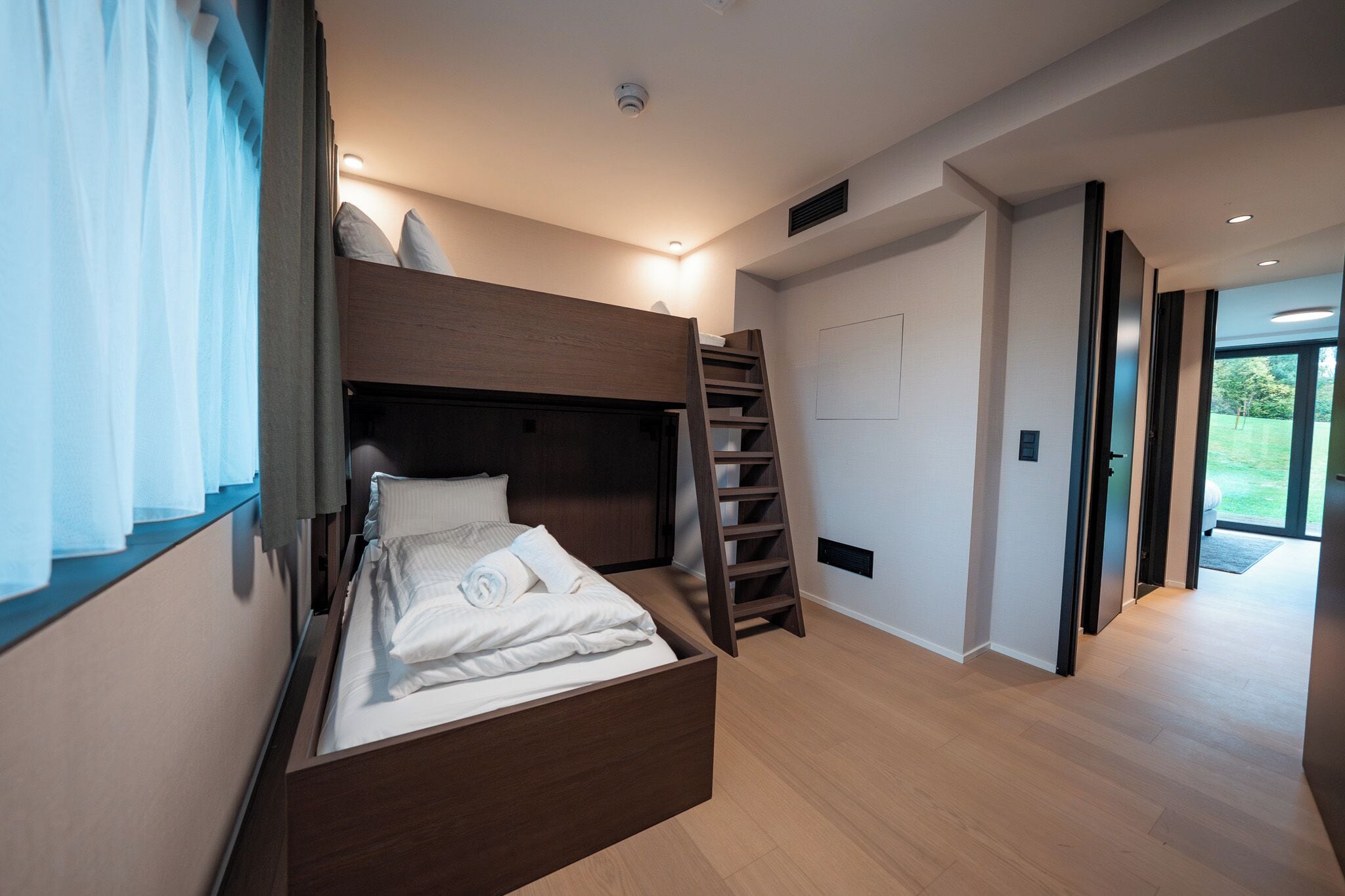 Un dormitorio moderno con una cama litera, una escalera de madera y vistas a un área verde al aire libre a través de una puerta.