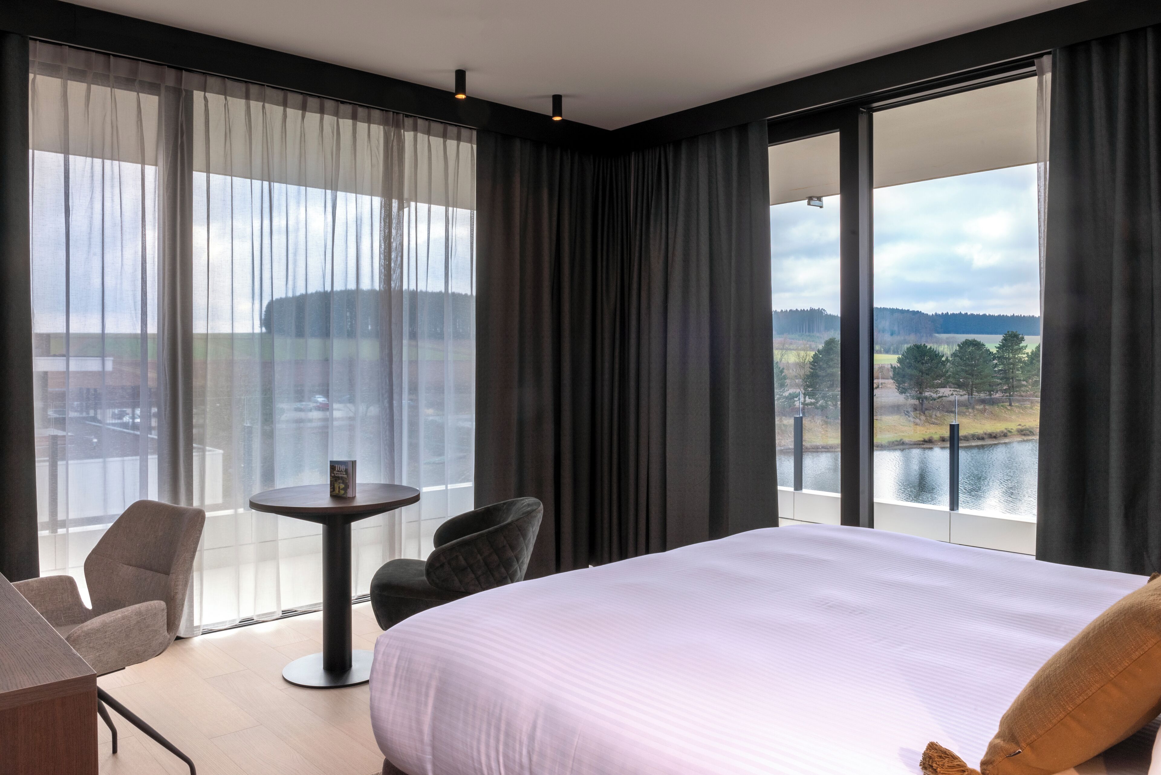 A hotel room with a large bed, a small table, two chairs, and a view of a lake and trees through large windows.