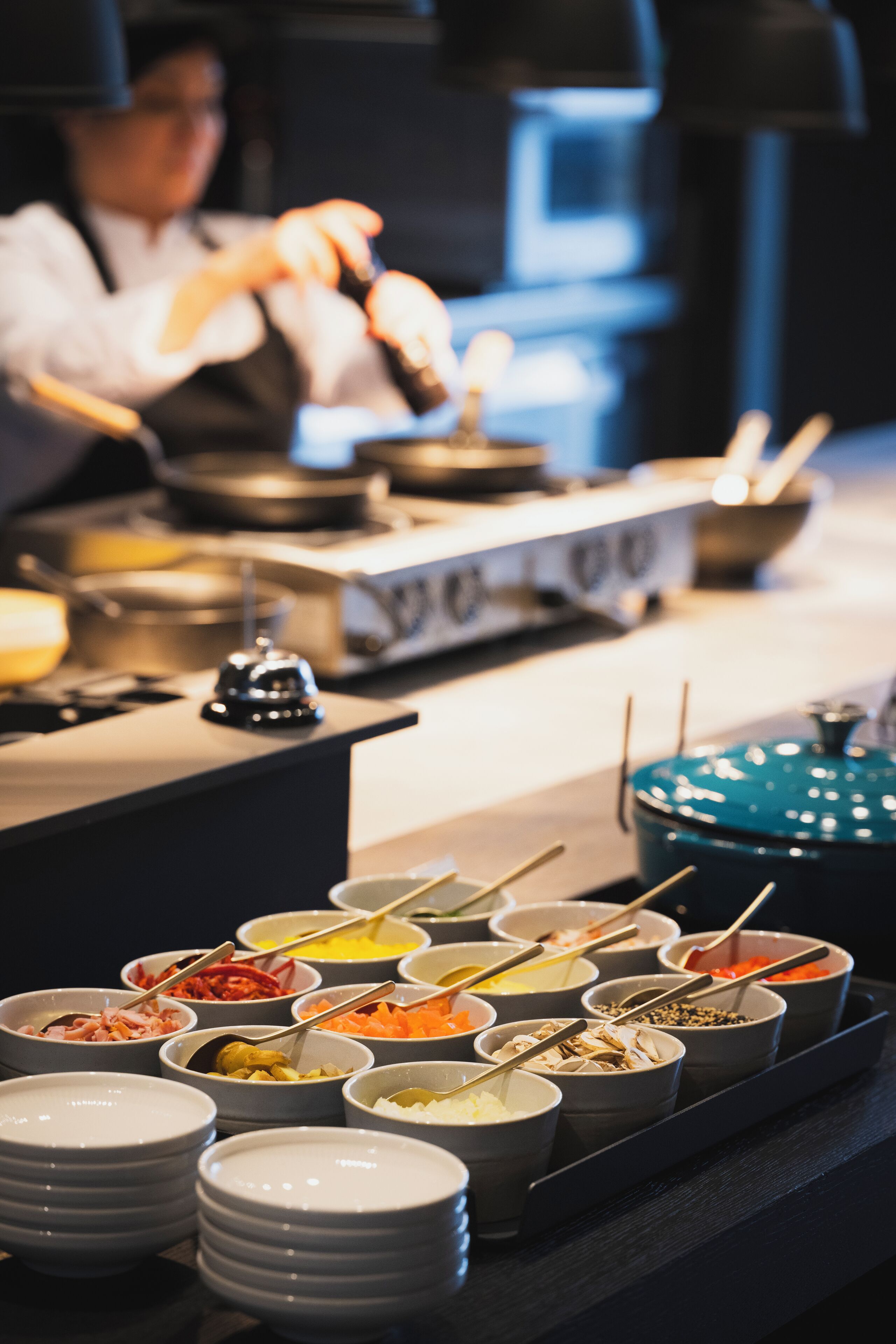 Un buffet con diversi piatti disposti in ciotole, uno chef sullo sfondo e una pentola blu sul bancone.