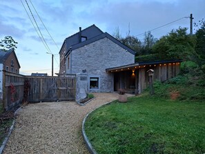 A stone building with a gravel driveway, a wooden fence, and a covered outdoor area with string lights. - La Tanière - Nature and Relaxation Gîte (Durbuy)