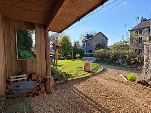 A wooden building with a covered patio, a bird feeder, a table with chairs, and a gravel path leading to a stone house. - La Tanière - Nature and Relaxation Gîte (Durbuy)