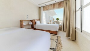 A hotel room with two beds, a wooden nightstand, a small table with a chair, and a large window with curtains. - New Spirit Dakhla (Dakhla)