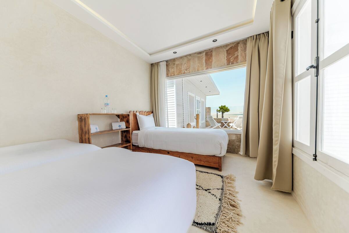 A hotel room with two beds, a wooden nightstand, a small table with a chair, and a large window with curtains.