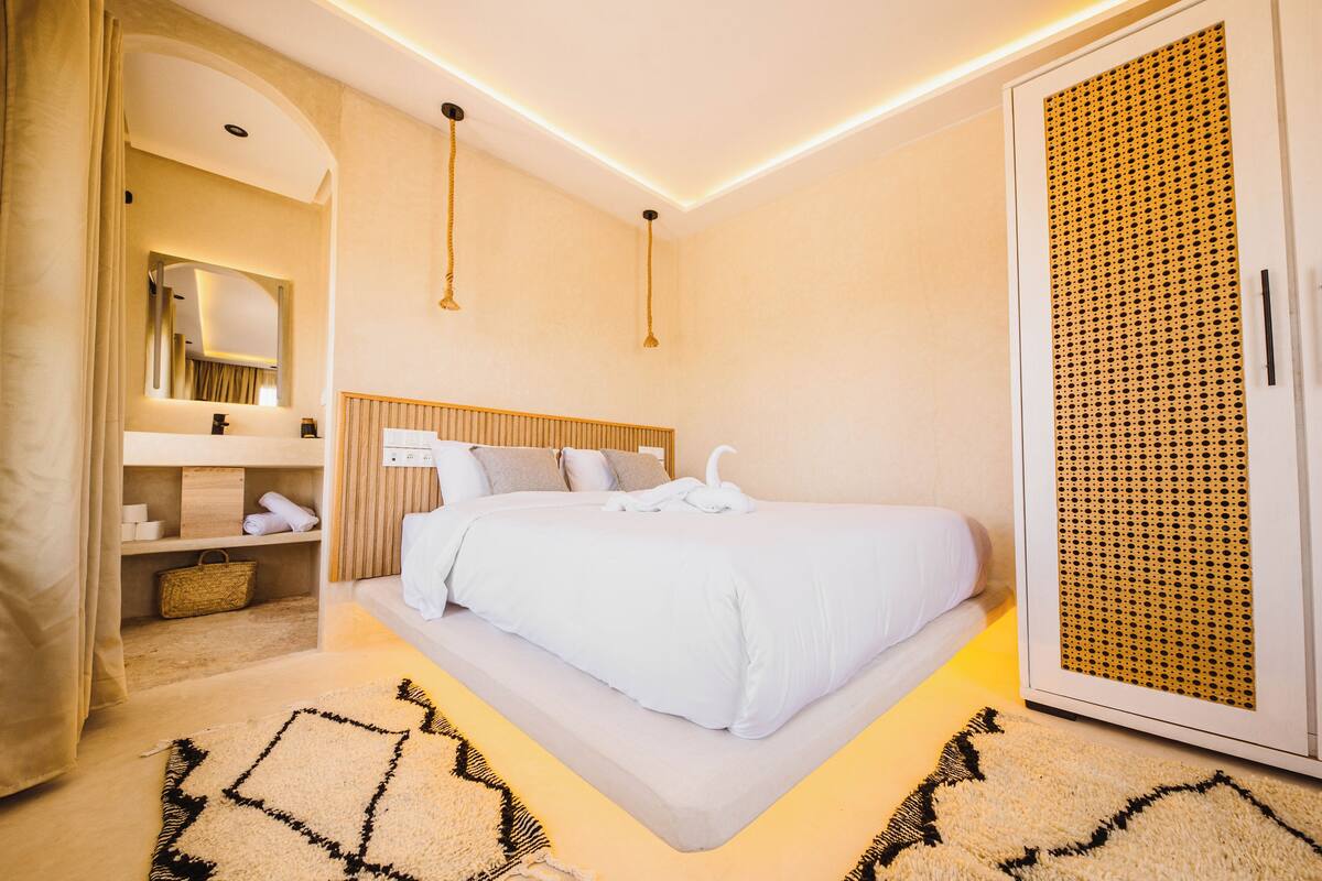 A modern hotel room with a large bed, a wooden headboard, a bedside table, a mirror, a wardrobe, and a patterned rug.