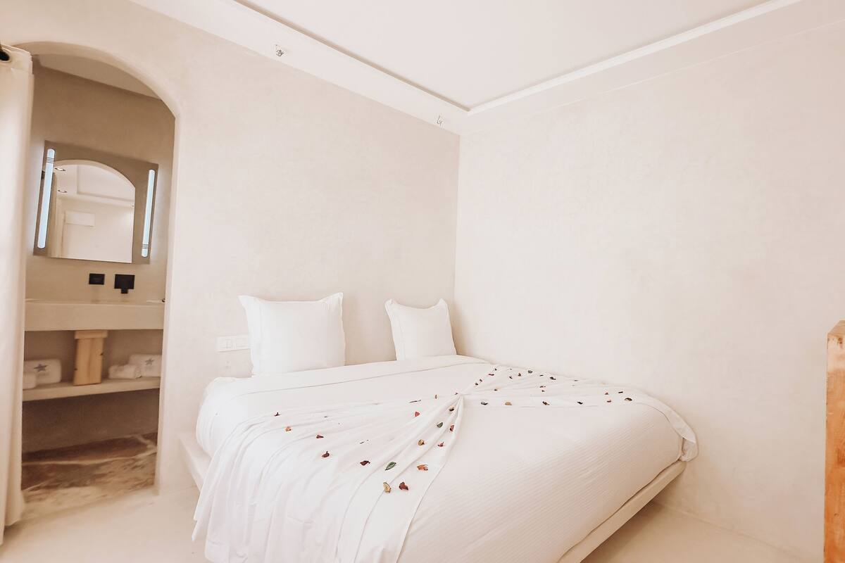 A minimalist bedroom with a bed, a wooden nightstand, a mirror, and a shelf with towels.