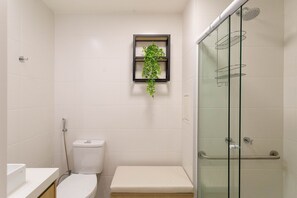 Een badkamer met toilet, een bankje, een glazen douchecabine en een plank met een plant.